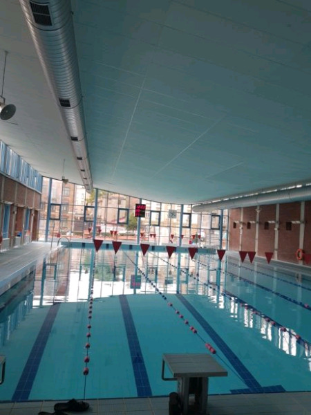 Wnętrze krytej pływalni z kilkoma torami oddzielonymi linami z czerwonymi bojami, widoczne okna z odbiciem zewnętrznego otoczenia, na końcu torów czerwone trójkątne flagi.