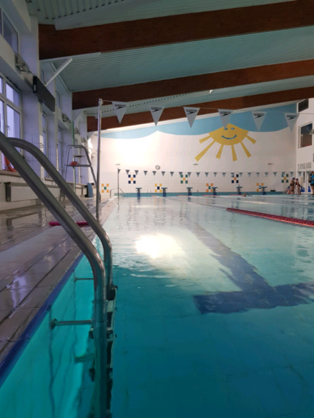 Kryty basen z drabinką wejściową, widoczne tory pływackie i dekoracja ścienna z motywem słońca.