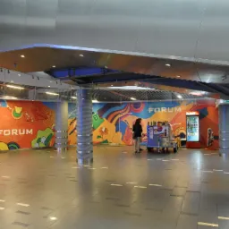 Wnętrze centrum handlowego Forum z kolorowymi grafikami na ścianach, widoczne filary z metalowymi zdobieniami, automat z napojami i wózek z przekąskami.