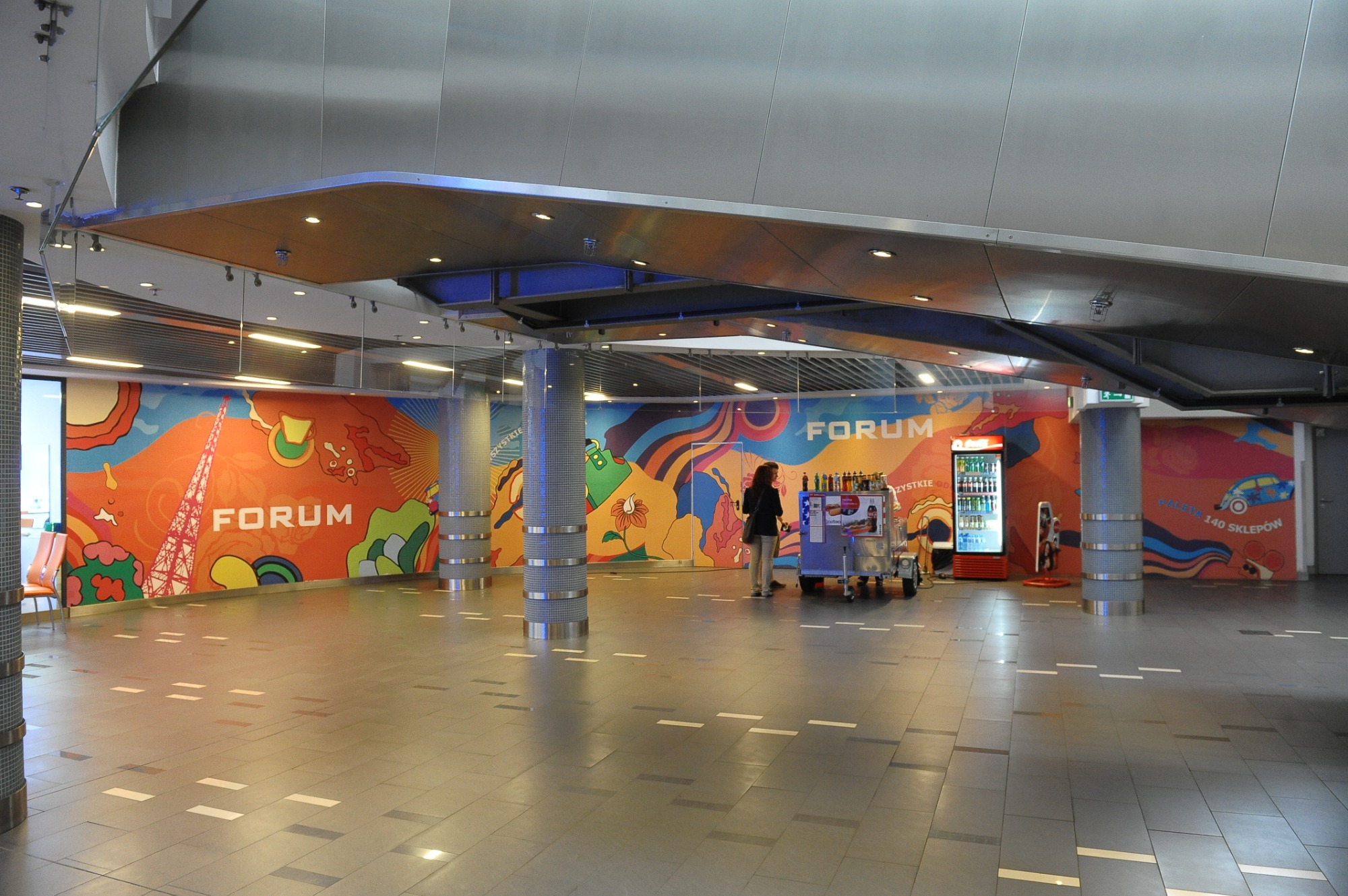 Wnętrze centrum handlowego Forum z kolorowymi grafikami na ścianach, widoczne filary z metalowymi zdobieniami, automat z napojami i wózek z przekąskami.