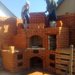 PEGAS Sp. z o.o. - Budowa zewnętrznego pieca z cegły: dwóch mężczyzn w roboczych ubraniach muruje konstrukcję z cegieł, widoczne metalowe wiadra, paleta z cegłami oraz łukowe otwory w piecu.