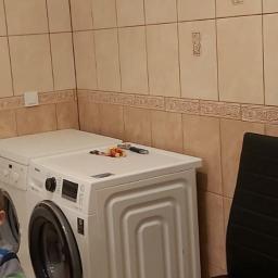 PEGAS Sp. z o.o. - Dwie białe pralki ustawione obok siebie w łazience z beżowymi płytkami, na jednej z nich leżą narzędzia i zabawki, obok stoi czarne krzesło, w kadrze widoczny fragment dziecka.