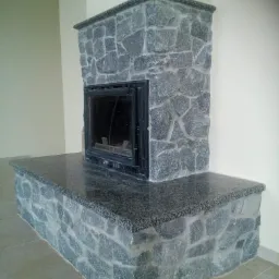 Obróbka kamieniarska kominka z szarego granitu, z widoczną fakturą kamienia i czarnym wkładem kominkowym.