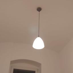 PEGAS Sp. z o.o. - Biała lampa wisząca w kształcie łzy oświetla kremowe ściany i sufit z delikatnym łukiem, widoczne okno z łukowatym nadprożem w tle.