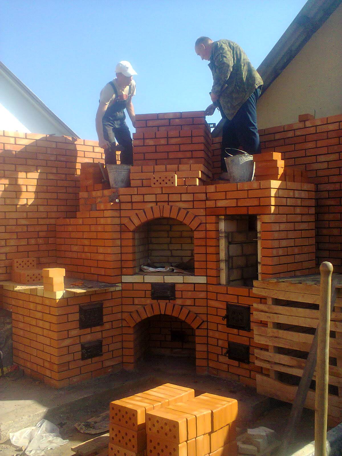 Budowa zewnętrznego pieca z cegły: dwóch mężczyzn w roboczych ubraniach muruje konstrukcję z cegieł, widoczne metalowe wiadra, paleta z cegłami oraz łukowe otwory w piecu.