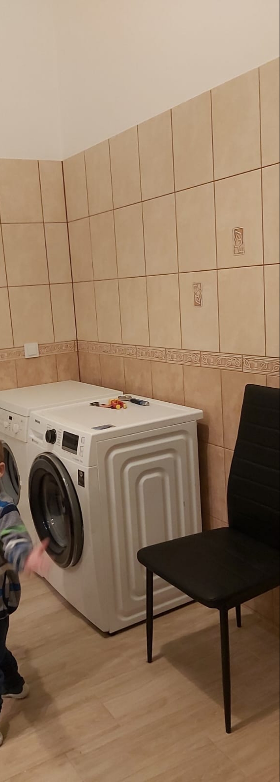 Dwie białe pralki ustawione obok siebie w łazience z beżowymi płytkami, na jednej z nich leżą narzędzia i zabawki, obok stoi czarne krzesło, w kadrze widoczny fragment dziecka.
