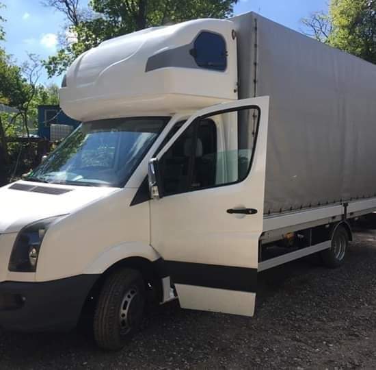 Biały samochód dostawczy z otwartymi drzwiami na żwirowym podłożu, widoczny fragment lasu w tle. Charakterystyczna zabudowa nad kabiną kierowcy.