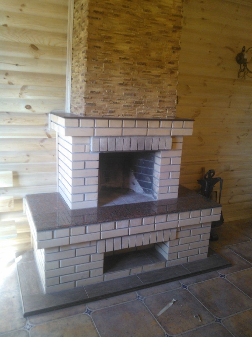 Murowany kominek z beżowych cegieł z blatem z brązowego granitu, otoczony drewnianą boazerią i kamienną okładziną komina, na podłodze brązowe płytki z dekoracyjnym wzorem.