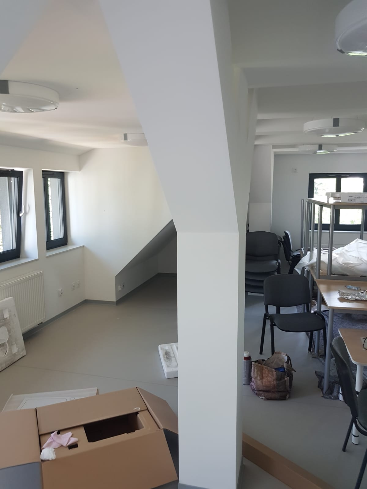 Wnętrze biura w trakcie odświeżania: świeżo pomalowane białe ściany, widoczny karton z akcesoriami, krzesła biurowe i stoliki w tle.