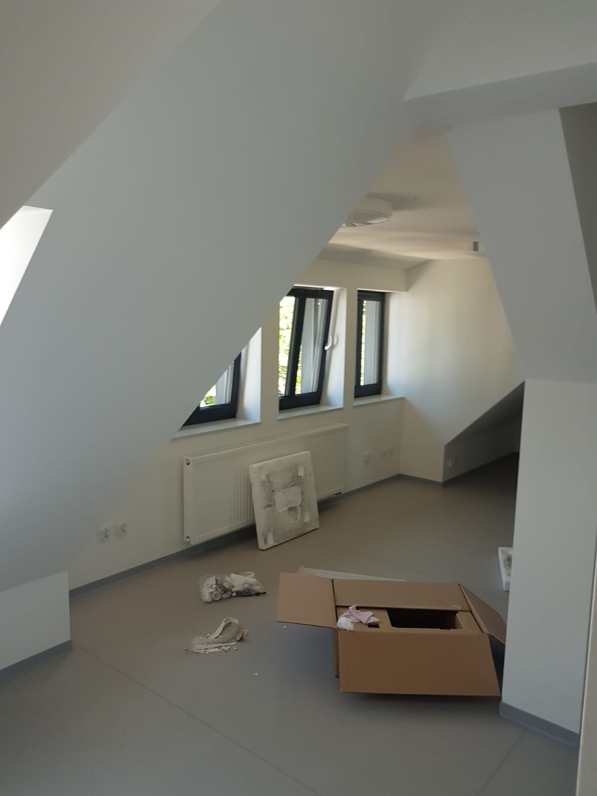 Poddasze po remoncie, ściany i sufit pomalowane na biało, szare panele na podłodze, okna w czarnych ramach, grzejniki na ścianach, karton na podłodze, widoczne ślady po pracy.
