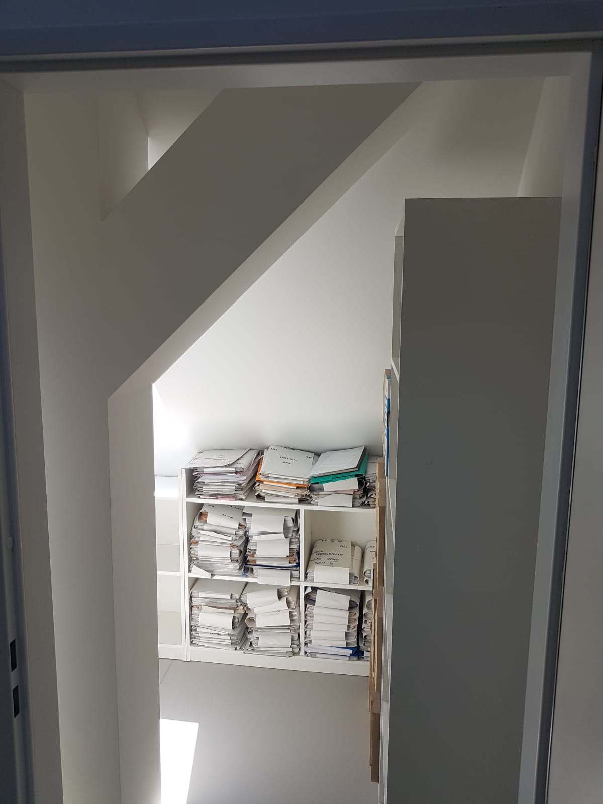 Biały regał z półkami wypełnionymi stertami dokumentów i segregatorów w pomieszczeniu ze skośnym sufitem, widoczne przez framugę drzwi.