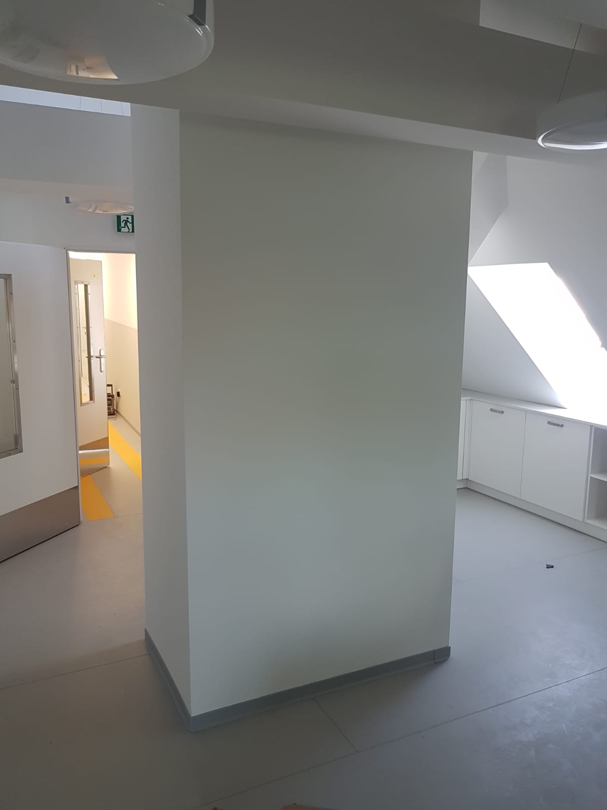 Świeżo pomalowane na jasno szaro ściany w biurze, widoczny fragment korytarza z żółtymi pasami na podłodze i białe szafki w tle.