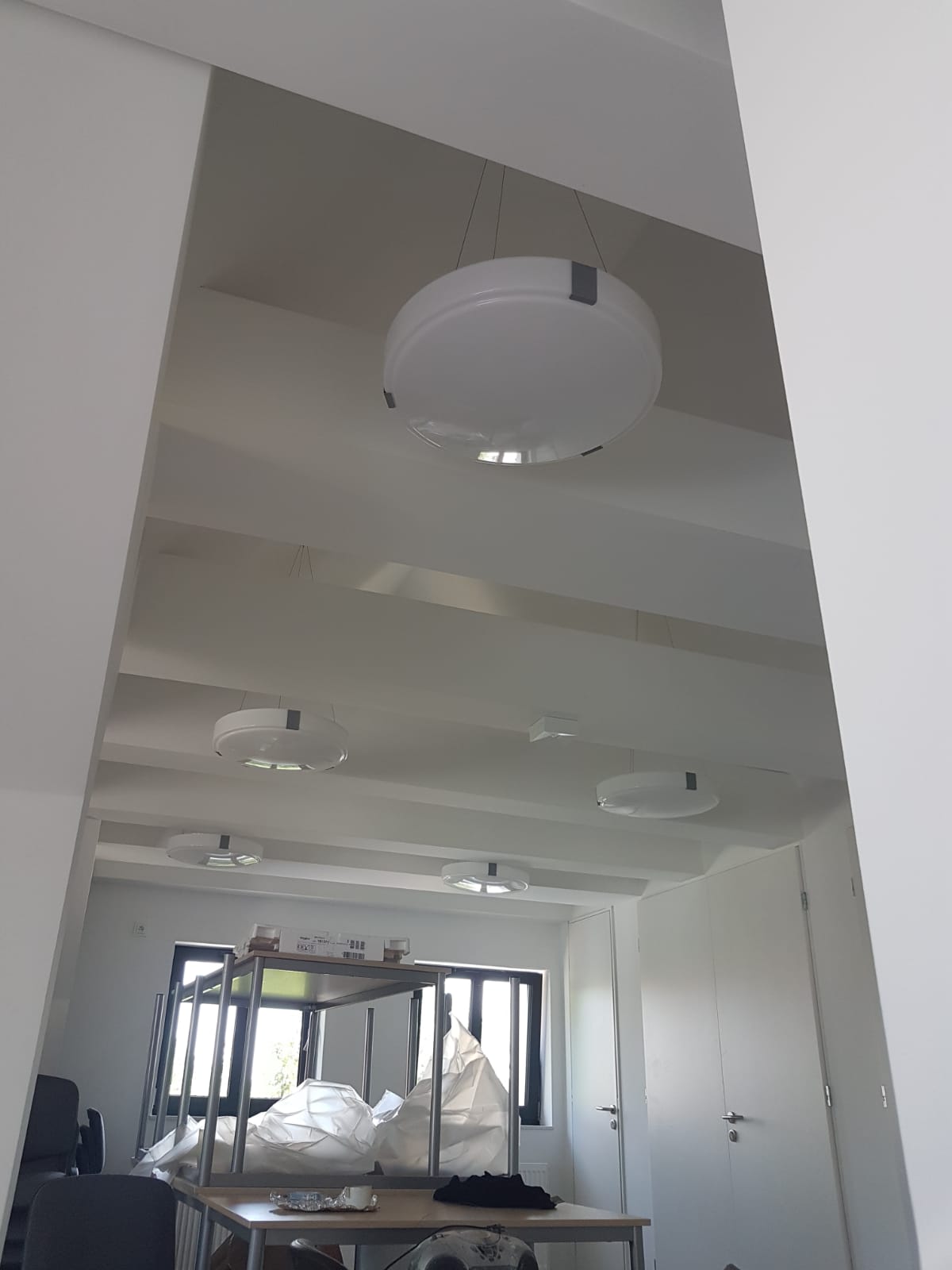 Białe, okrągłe lampy sufitowe zawieszone na cienkich linkach w jasnym biurze z białymi ścianami i belkami sufitowymi, widoczne meble biurowe w trakcie remontu, częściowo zakryte folią.