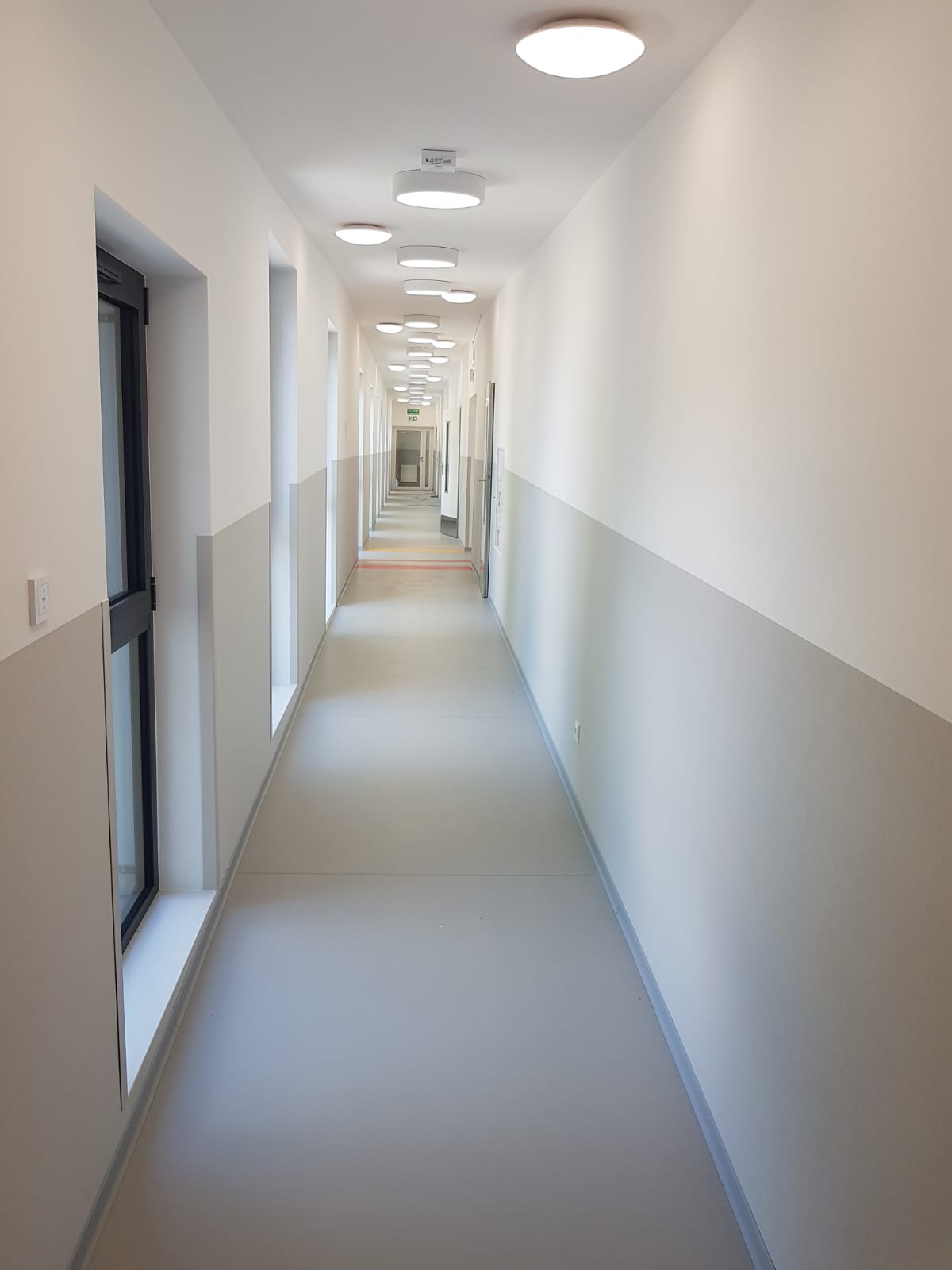Długi, pusty korytarz w biurze lub budynku użyteczności publicznej, z białymi ścianami z szarym pasem na dole, równo rozmieszczonymi okrągłymi lampami sufitowymi i szarą podłogą.