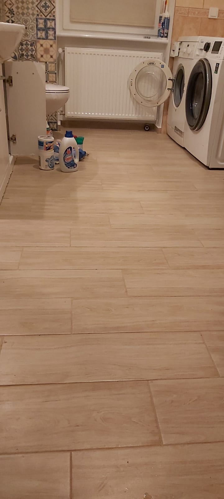 Wykończona łazienka w Szczecinie z podłogą wyłożoną jasnymi, drewnopodobnymi płytkami, widoczna pralka z otwartymi drzwiczkami, biała szafka pod umywalką oraz kolorowe płytki dekoracyjne na ścianie.