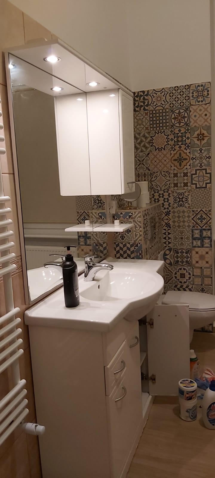 Umywalka z szafką w łazience z wzorzystą ścianą, widoczne otwarte drzwiczki szafki i akcesoria łazienkowe, czarny dozownik mydła na blacie.