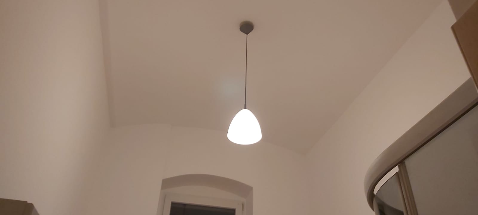 Biała lampa wisząca w kształcie łzy oświetla kremowe ściany i sufit z delikatnym łukiem, widoczne okno z łukowatym nadprożem w tle.