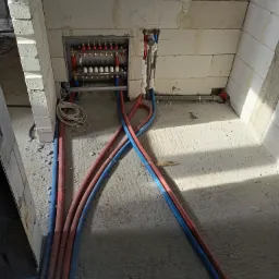 Rozdzielacz ogrzewania podłogowego zamontowany w ścianie z bloczków, z podłączonymi rurami w kolorach czerwonym i niebieskim, ułożonymi na surowej posadzce betonowej.