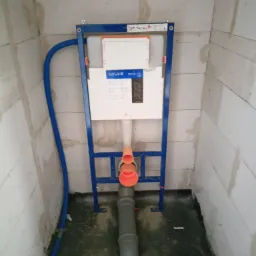 Stelaż podtynkowy Cersanit Tech Line z widocznymi rurami kanalizacyjnymi i dopływową niebieską rurą wodną, zamontowany w surowym pomieszczeniu z bloczków betonowych.