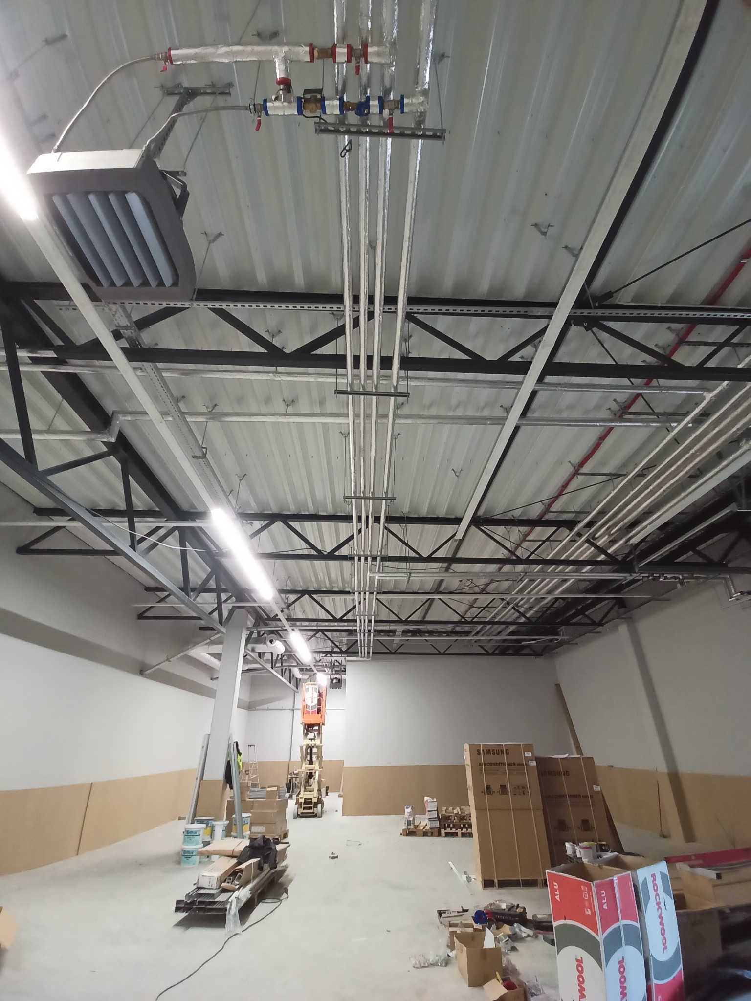 Wnętrze hali w trakcie instalacji systemów grzewczych i wentylacyjnych; widoczne rury, grzejnik przemysłowy podwieszony pod sufitem, materiały izolacyjne i podnośnik koszowy.