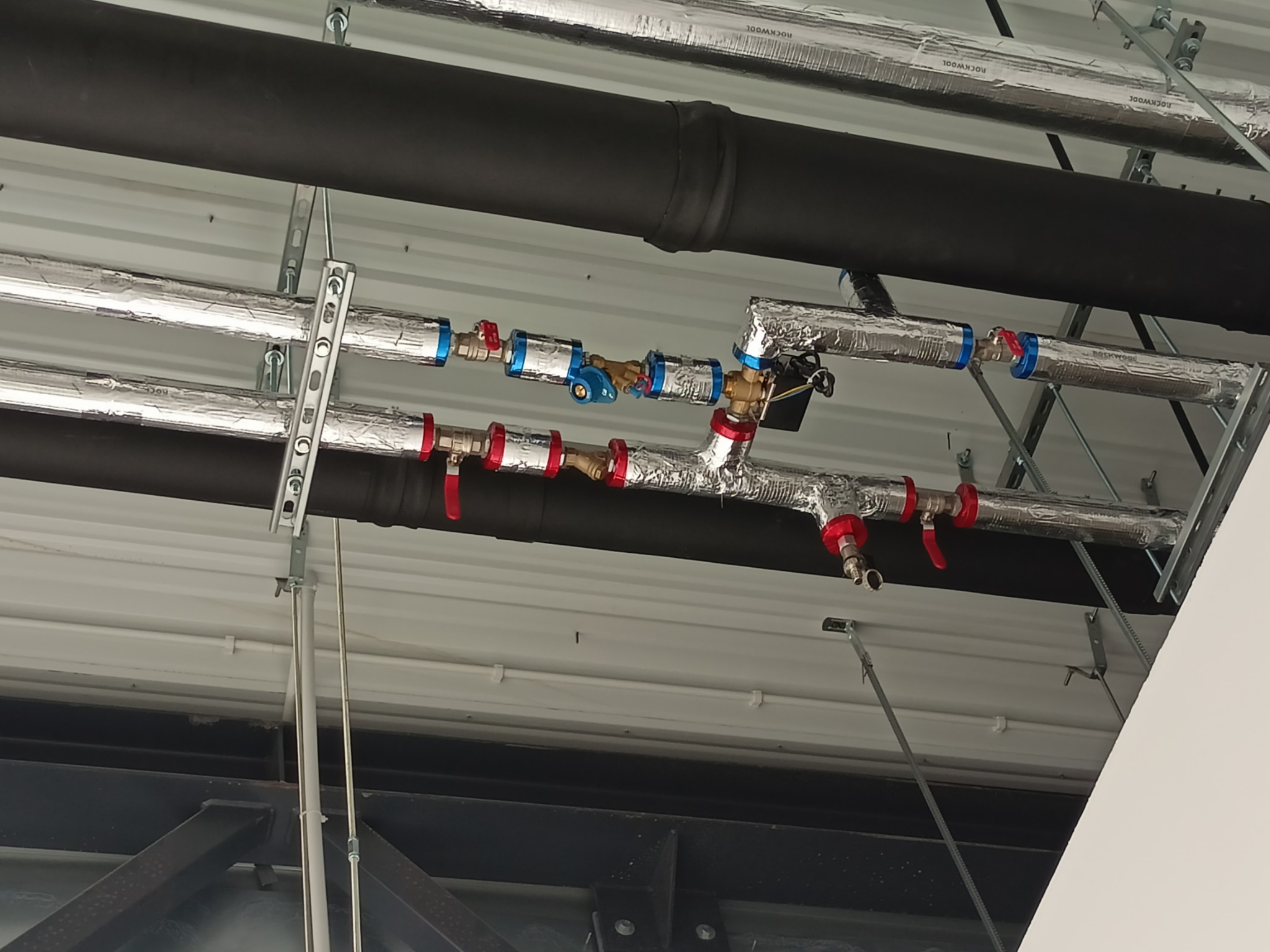 Widok instalacji grzewczej i hydraulicznej pod sufitem, rury z izolacją aluminiową, zawory z czerwonymi i niebieskimi pokrętłami, czarne rury izolowane termicznie, mocowania do sufitu.