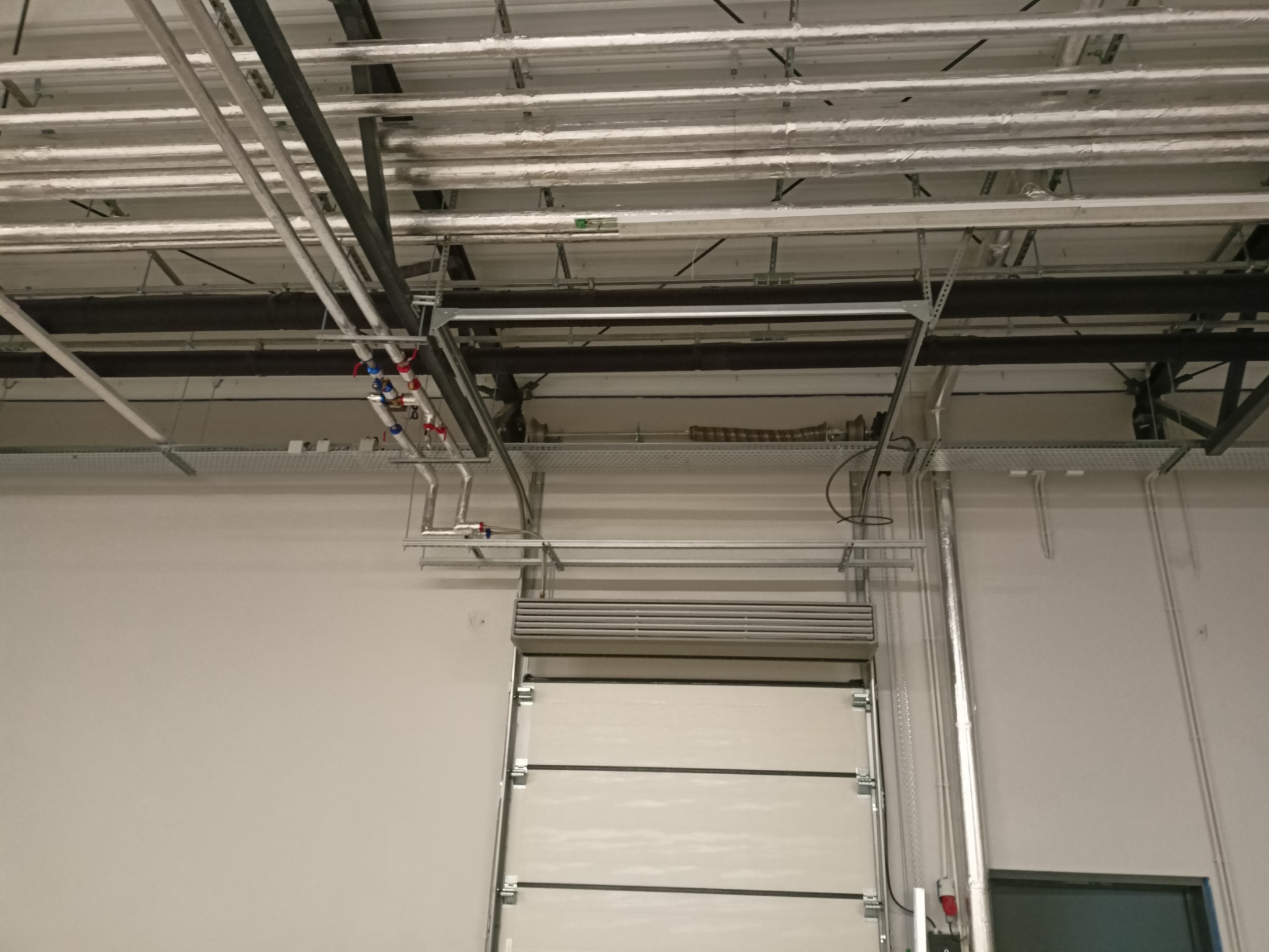 Widok instalacji rur i przewodów pod sufitem w hali przemysłowej z bramą segmentową w tle.