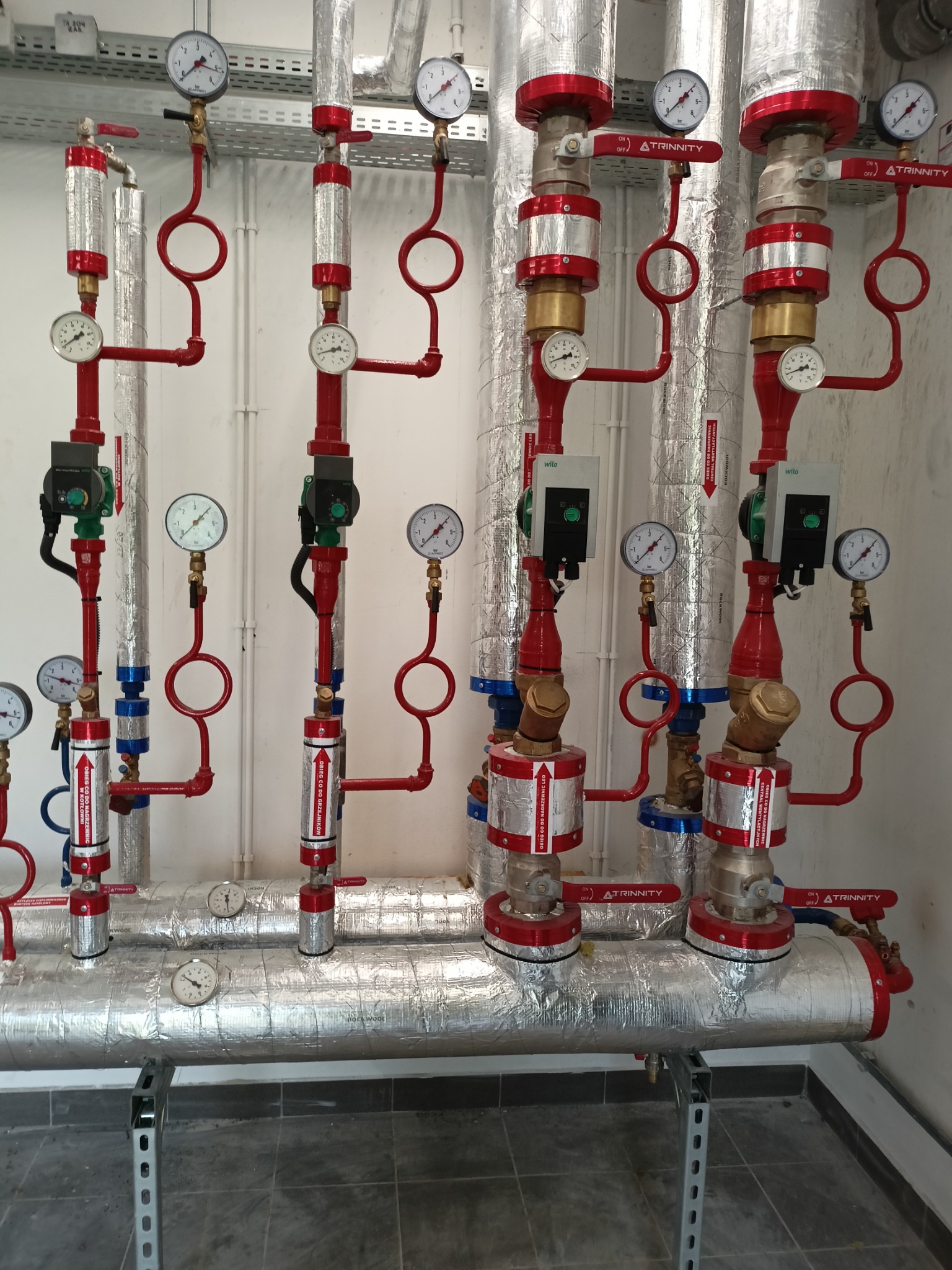 Złożona instalacja grzewcza z rur izolowanych folią aluminiową, manometrami, zaworami z czerwonymi uchwytami oznaczonymi 'ATRINNITY' i pompami obiegowymi Wilo, widoczna na tle białej ściany.