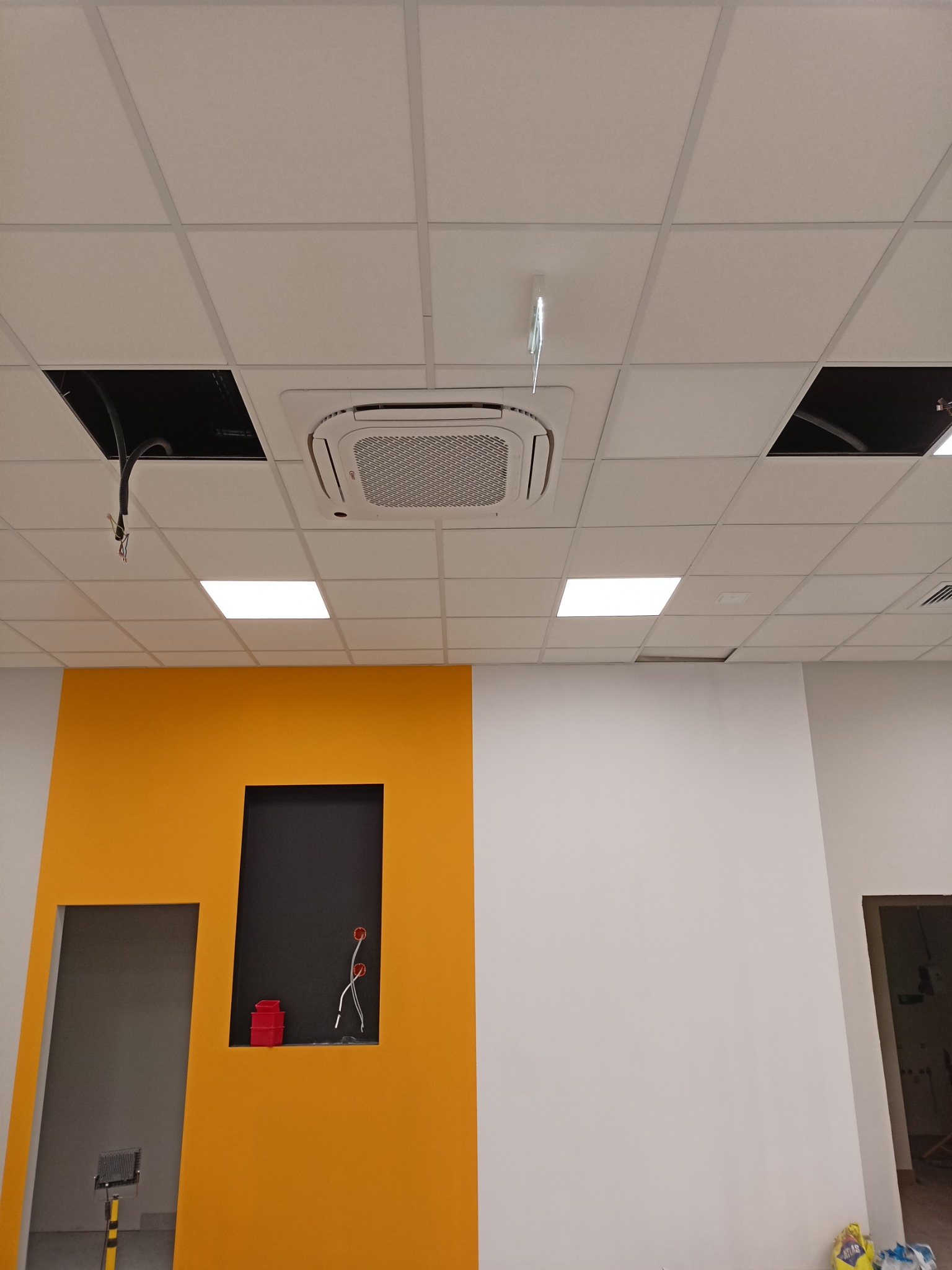 Sufit podwieszany z zamontowaną kasetą klimatyzacyjną, widoczne przewody w otworach, ściana pomalowana na żółto z czarnymi wnękami, w jednej z nich czerwone pojemniki i wystające przewody.