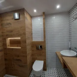 Nowoczesna łazienka z drewnopodobnymi płytkami na ścianach, białymi płytkami typu metro, wzorzystą podłogą, wiszącą toaletą i umywalką na drewnianym blacie.