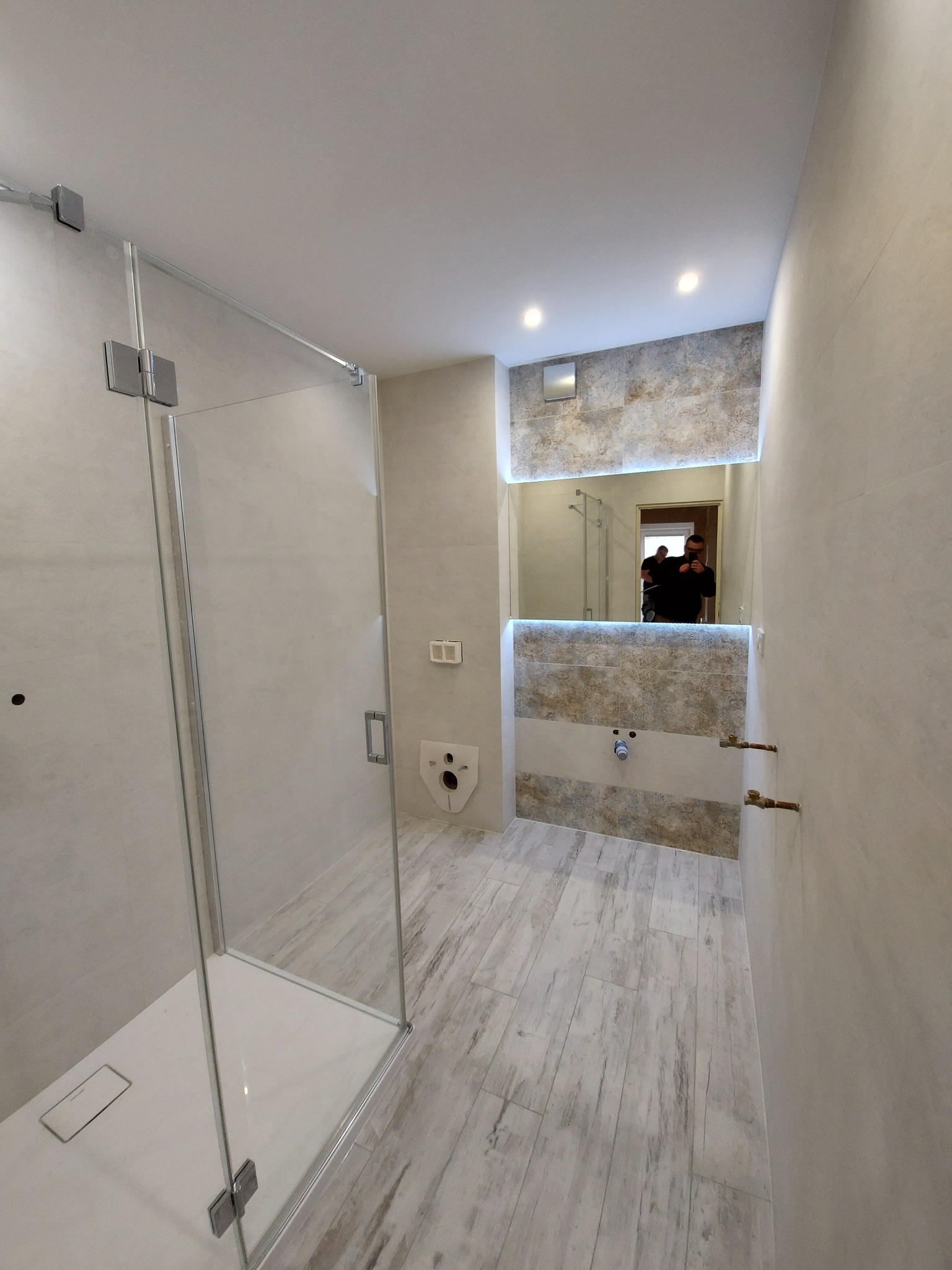 Nowoczesna łazienka z kabiną prysznicową typu walk-in, drewnopodobnymi płytkami na podłodze i lustrem z podświetleniem LED, widoczne przyłącza wodne.