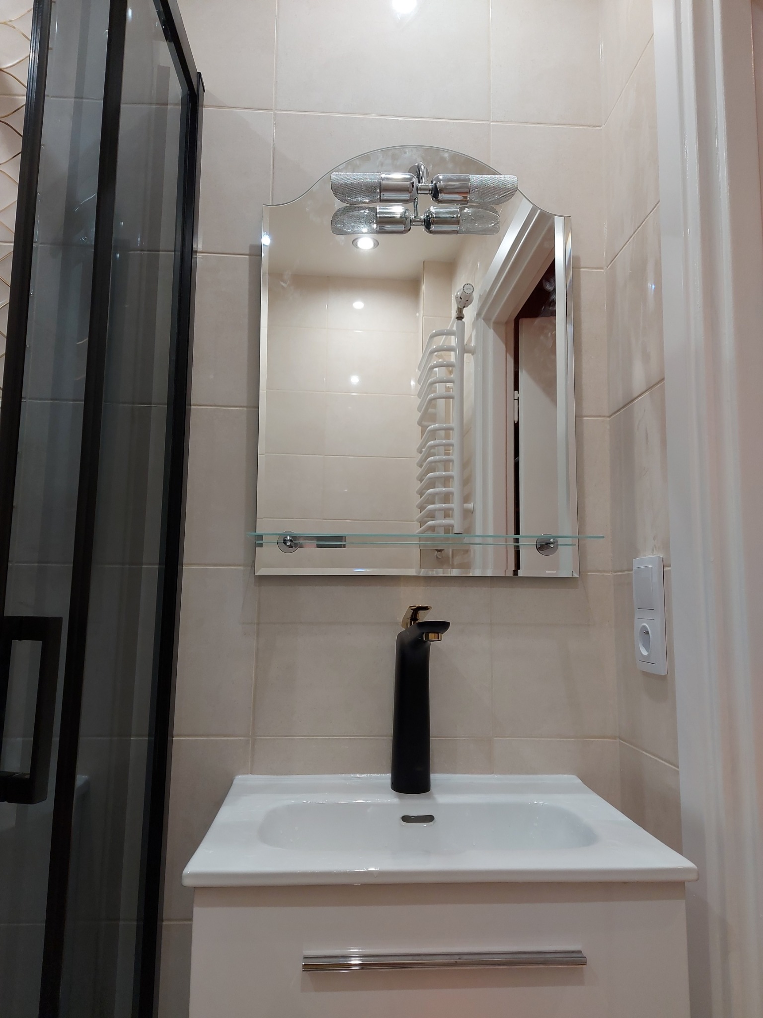 Nowoczesna łazienka z beżowymi płytkami, czarną armaturą i kabiną prysznicową z czarną ramą. Lustro z oświetleniem nad umywalką odbija grzejnik drabinkowy.
