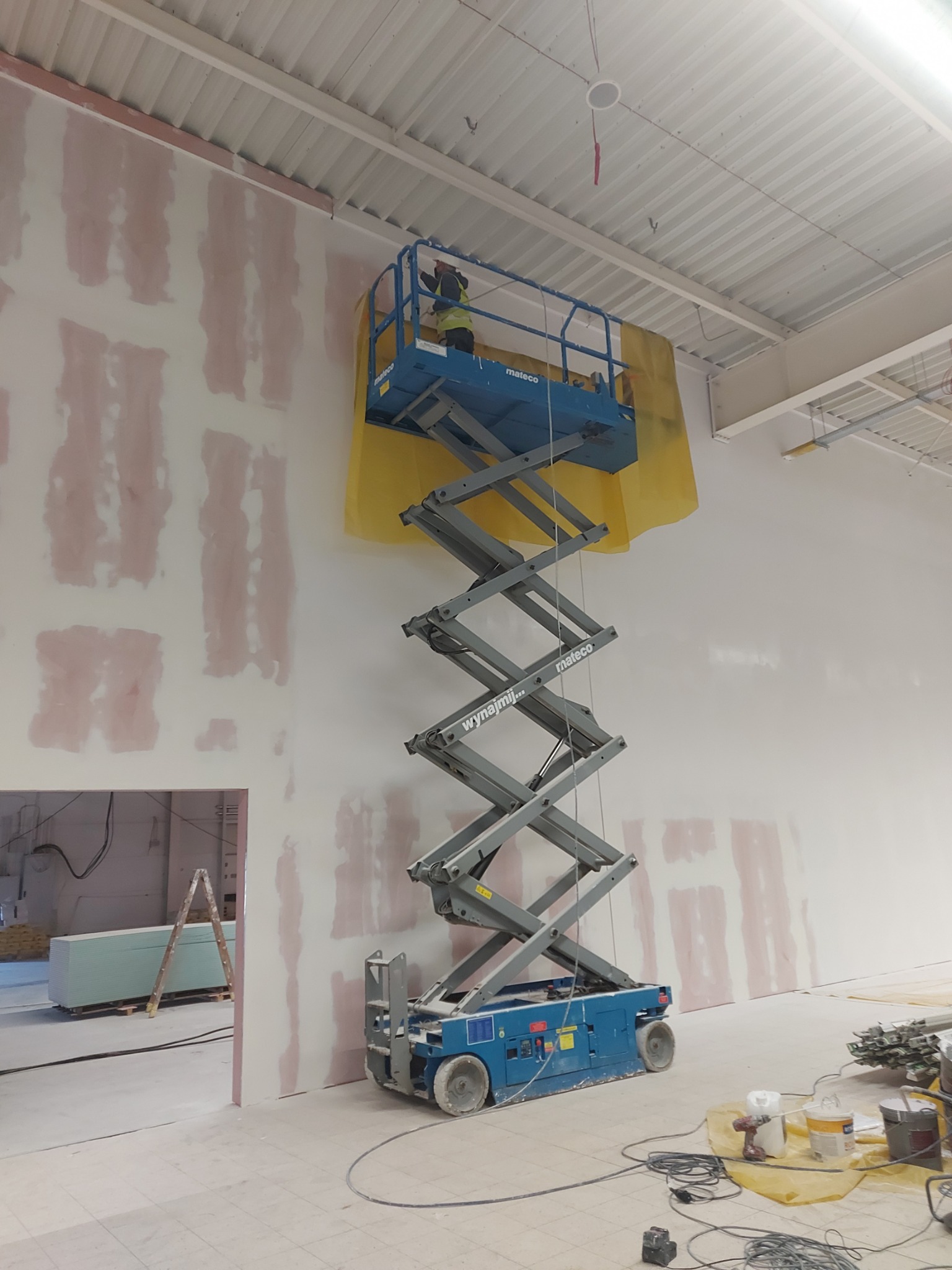 Pracownik na podnośniku nożycowym, zabezpieczonym folią, wykonuje prace na wysokości na ścianie z nałożonymi pasami szpachli, widoczne płyty kartonowo-gipsowe i drabina w tle.