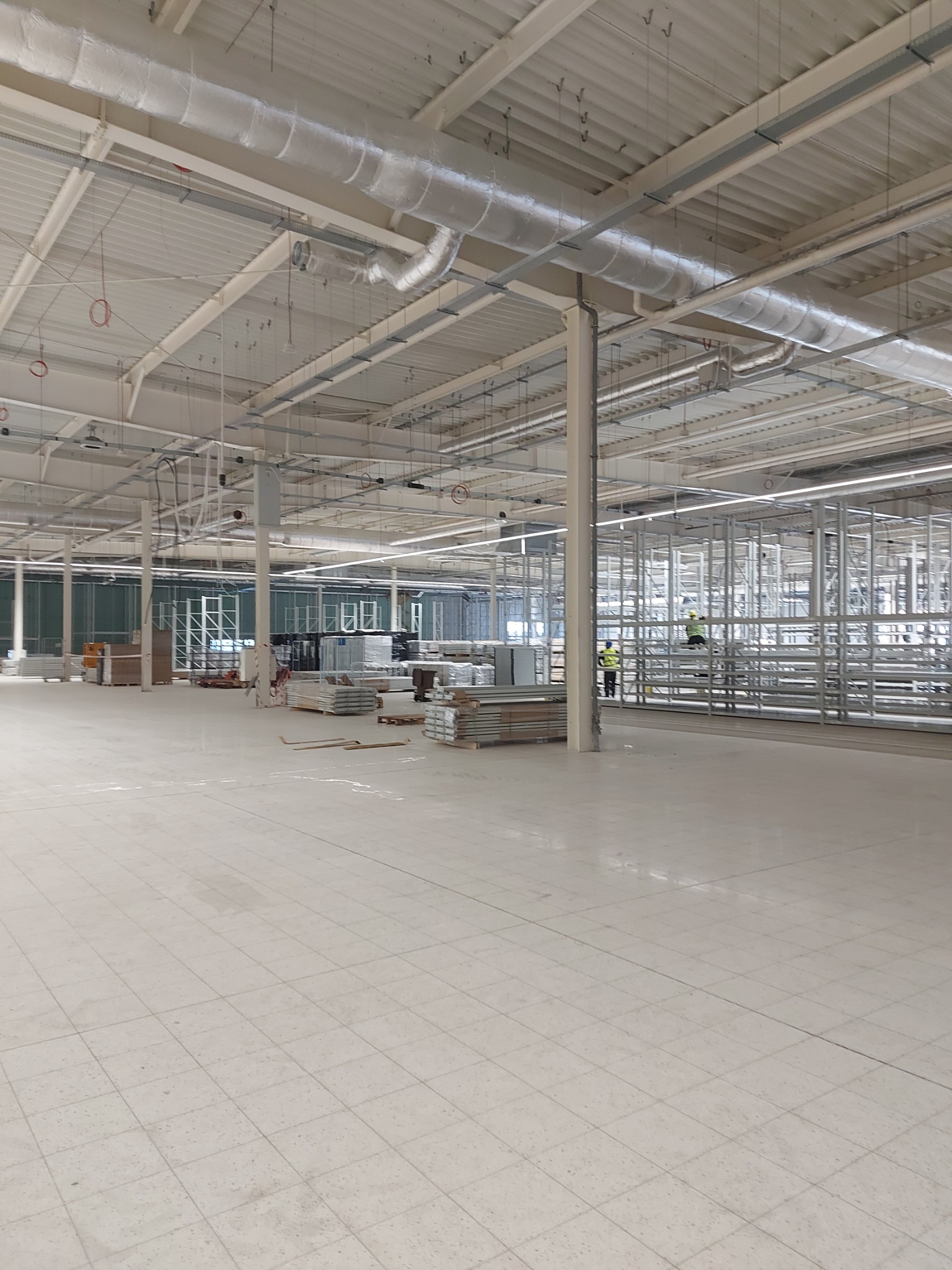 Przestronne wnętrze hali w trakcie prac wykończeniowych, widoczne metalowe konstrukcje ścian działowych, instalacje pod sufitem oraz pracownicy w oddali.