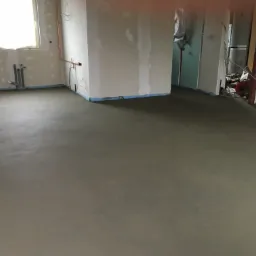 Świeżo wylana, gładka, szara posadzka w surowym wnętrzu z instalacjami hydraulicznymi przy ścianie i niezagruntowanymi ścianami przygotowanymi do malowania.