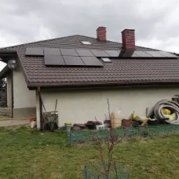 Brązowy dach z panelami słonecznymi na domu w Tomaszowie Mazowieckim, widok z trawnika z młodym drzewkiem w siatce ochronnej.