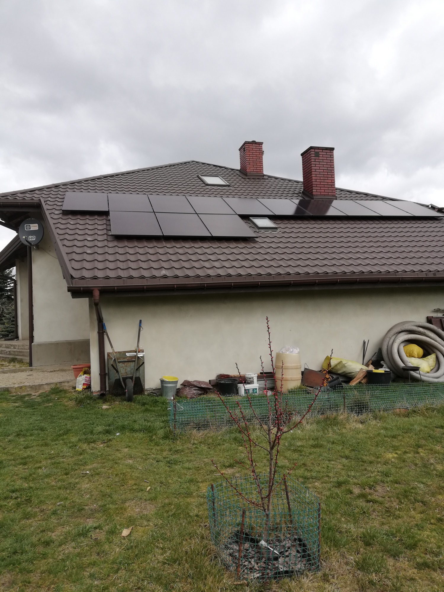 Brązowy dach z panelami słonecznymi na domu w Tomaszowie Mazowieckim, widok z trawnika z młodym drzewkiem w siatce ochronnej.