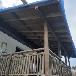 Drewniany taras z balustradą w trakcie budowy, widoczna drabina oparta o ścianę budynku, niebieska plandeka zabezpiecza fragment dachu.