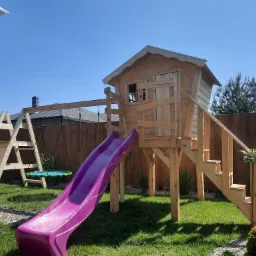 Drewniany plac zabaw dla dzieci z domkiem na platformie, schodami, zjeżdżalnią w kolorze fuksji i huśtawką, ustawiony na trawniku w słoneczny dzień.