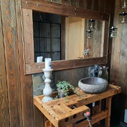 Rustykalna łazienka z drewnianymi ścianami, lustrem w drewnianej ramie z metalowymi nitami, kamienną umywalką na starym stole warsztatowym, świecznikiem i rośliną w puszce, oświetlona wiszącymi...