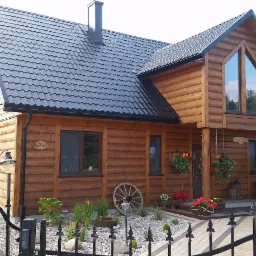 Dwukondygnacyjny dom z bali z czarnym dachem, widoczny zza czarnego metalowego ogrodzenia z kamiennym podłożem i dekoracyjnym ogrodem.