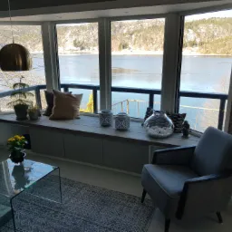 Nowoczesny salon z dużym oknem wykuszowym, drewnianym siedziskiem z szufladami, dekoracjami, widokiem na jezioro i zaśnieżone wzgórza, szary fotel i szklany stolik kawowy.