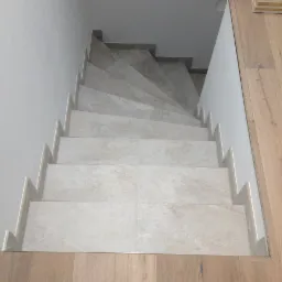 Schody wyłożone jasnymi płytkami imitującymi kamień, z widocznymi listwami przypodłogowymi i drewnianą podłogą na dole.