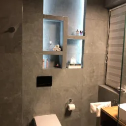 Nowoczesna łazienka z szarymi płytkami imitującymi kamień, podświetlanymi wnękami na kosmetyki i akcesoria, wiszącą toaletą oraz drewnianą szafką pod umywalką.