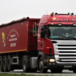 Czerwona ciężarówka Scania z naczepą, z logo firmy Speedtrans i informacją o transporcie piasku, żwiru i zboża, widoczna na drodze w pochmurny dzień, z osobowym samochodem w tle.