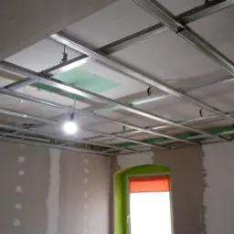 Sufit podwieszany z metalową konstrukcją i częściowo zamontowanymi płytami gipsowymi, widoczne fragmenty ścian po szpachlowaniu, okno z pomarańczową roletą i zieloną ramą.