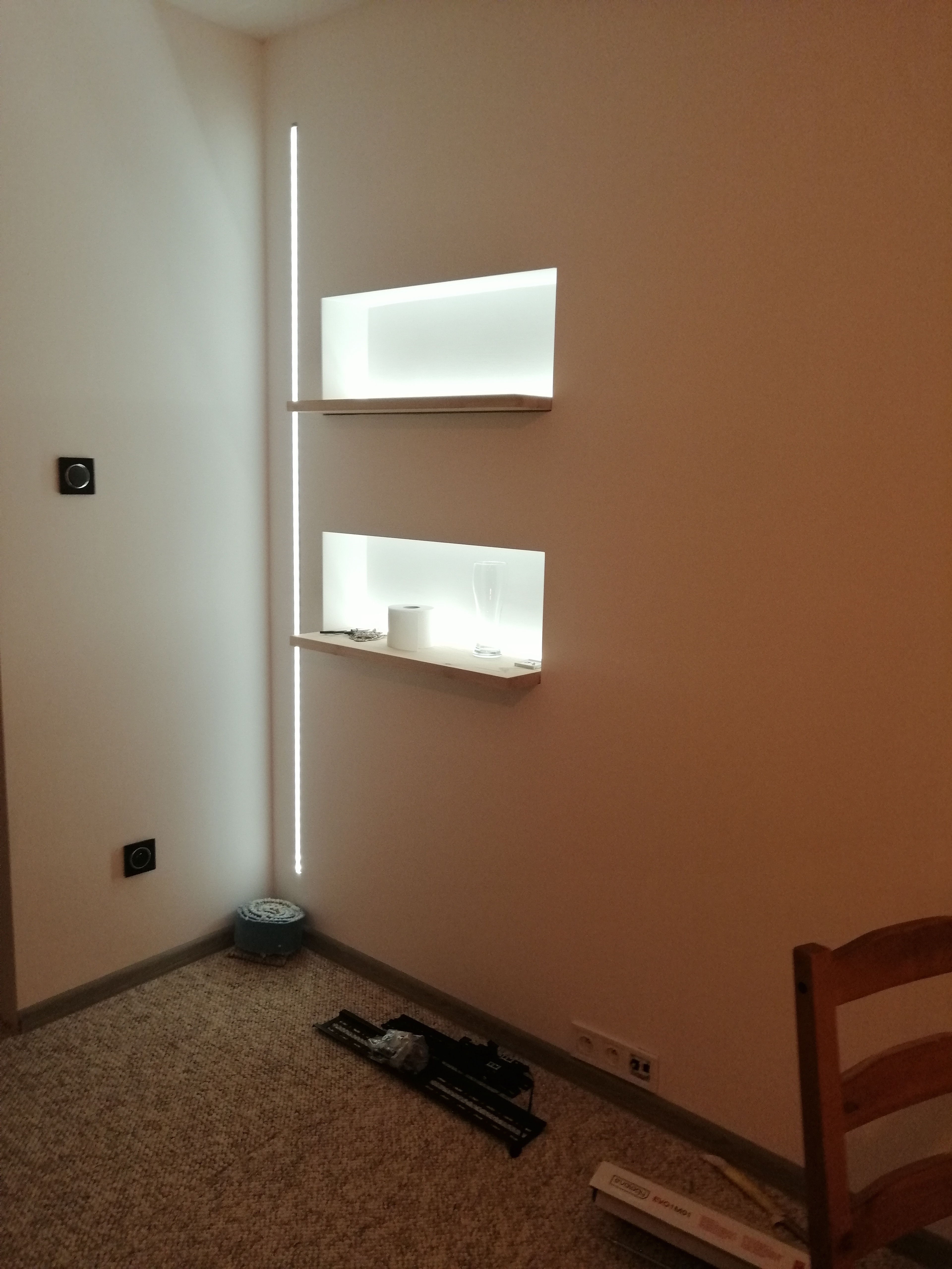 Nowoczesne, podświetlane wnęki w ścianie z półkami, na których leżą dekoracje. Na podłodze widoczne narzędzia i materiały budowlane, sugerujące prace wykończeniowe w toku.