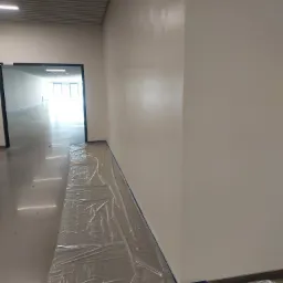 Świeżo pomalowana, jasna ściana w korytarzu, zabezpieczona folią i taśmą malarską, widoczne delikatne smugi po wałku.