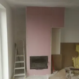 Wnętrze w trakcie prac wykończeniowych: obudowa kominka z płyt kartonowo-gipsowych, widoczna drabina aluminiowa, poziomica i fragmenty materiałów budowlanych.