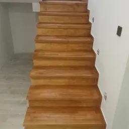 Nowe drewniane schody wewnętrzne z jasnego drewna, widoczne z dołu, z białymi ścianami po bokach, na których widać wystające kable elektryczne.