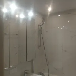 Nowoczesna łazienka z marmurowymi płytkami, zabudowaną szafką z lustrem nad umywalką i prysznicem ze szklaną ścianką.